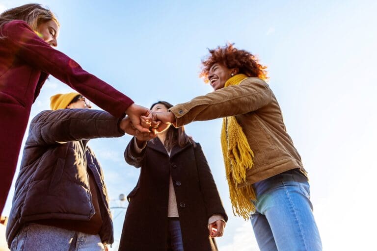 A dynamic low-angle shot capturing a group of friends with different ethnic backgrounds giving a fist bump in a circle under a clear blue sky. The sun is casting a warm glow on their happy faces, highlighting a woman with curly hair wearing a mustard yellow scarf who is laughing joyfully. The image conveys a sense of unity and camaraderie among the group.