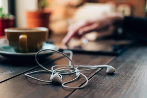 White earphones lying tangled on a wooden table next to a yellow cup of coffee, with a blurred background showing a hand interacting with a tablet and some potted plants.